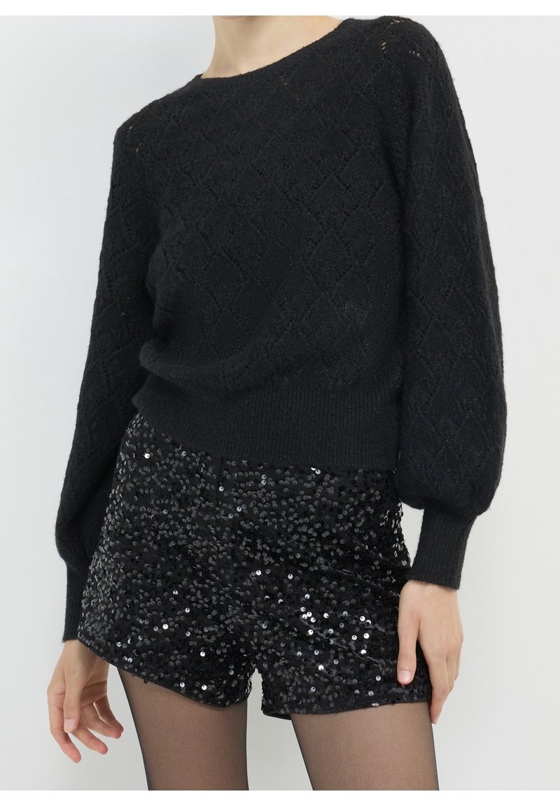 Femme portant un pull texturé noir à manches longues et un short noir à sequins sur des collants noirs transparents.
