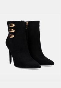 Botas de tobillo de ante negro con punta afilada, tacón de aguja y tres detalles de hardware dorado en el lateral. Cierre con cremallera lateral.