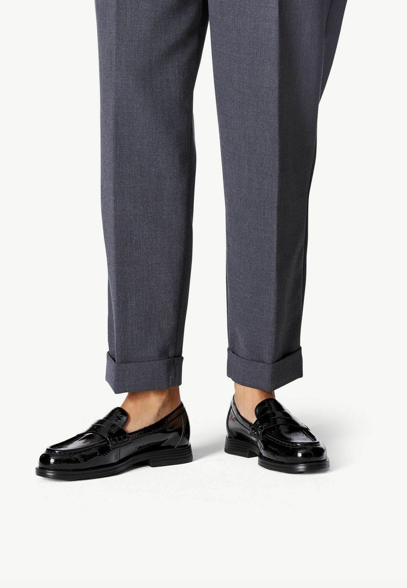 Persona vestida con pantalones grises de corte a medida con dobladillo y zapatos mocasines de cuero negro brillante, de pie sobre un fondo blanco.