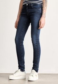 Úzke rifle z tmavomodrej džínoviny s klasickým päťvreckovým dizajnom, jemným prešívaním a stredným pásom. Kombinované s bielymi teniskami na platforme.