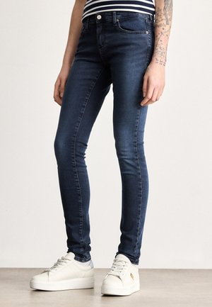 Úzke rifle z tmavomodrej džínoviny s klasickým päťvreckovým dizajnom, jemným prešívaním a stredným pásom. Kombinované s bielymi teniskami na platforme.