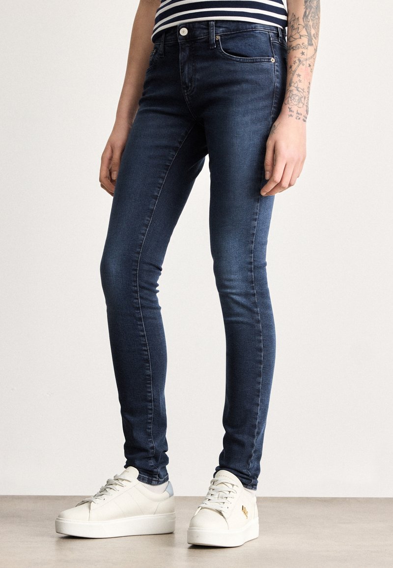 Úzke rifle z tmavomodrej džínoviny s klasickým päťvreckovým dizajnom, jemným prešívaním a stredným pásom. Kombinované s bielymi teniskami na platforme.