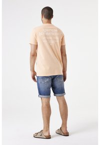 Camiseta de manga corta color durazno claro con texto blanco en la parte de atrás, combinada con pantalones cortos de mezclilla remangados y sandalias beige. Diseño simple y casual.