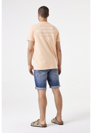 Lichtperzikkleurig T-shirt met korte mouwen en witte tekst op de achterkant, gecombineerd met opgerolde denim shorts en beige sandalen. Eenvoudig, casual ontwerp.