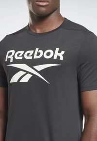 Camiseta de algodón gris con un gran logo blanco de Reebok y diseño gráfico. Mangas cortas, cuello redondo, textura suave.