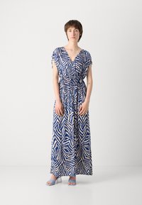 Robe maxi bleu marine avec un motif rayé zèbre crème, décolleté en V, manches courtes et taille élastique. Le modèle porte des sandales à talons bleues.