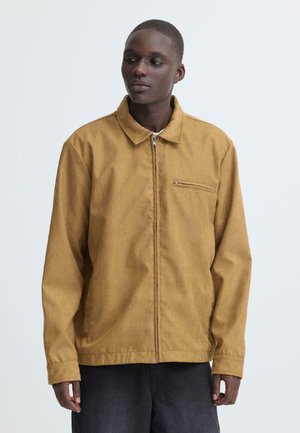 Man draagt een beige jack met ritssluiting en een klein borstvak met rits, staande tegen een effen lichtgrijze achtergrond.