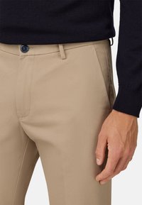 Beigās bikses ar gludu tekstūru, kas aprīkotas ar metāla pogu aizdari un sānu kabatām, saskaņotas ar tumši zilu džemperi.