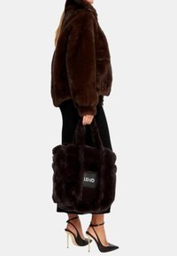 Borsa in pelliccia sintetica marrone con manici corti e pelosi e forma rettangolare. Presenta un'etichetta di marca visibile. Indossata con una giacca marrone coordinata e scarpe con tacco alto.