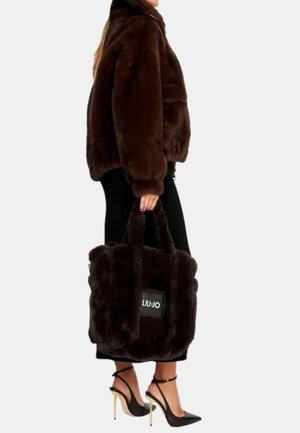 Borsa in pelliccia sintetica marrone con manici corti e pelosi e forma rettangolare. Presenta un'etichetta di marca visibile. Indossata con una giacca marrone coordinata e scarpe con tacco alto.