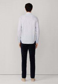 Hombre con cabello rizado que lleva una camisa de manga larga a cuadros claros, pantalones oscuros y zapatos beige, de pie de espaldas sobre un fondo liso.