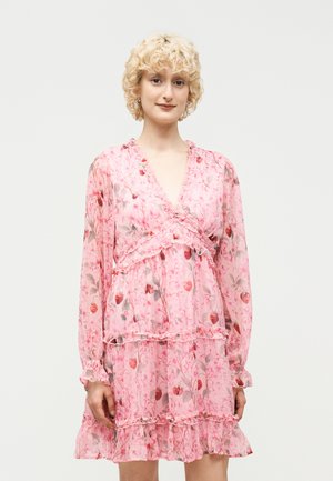 Person med kort krøllet blondt hår iført en lyserød blomsterprintet langærmet kjole med flæsetrin og V-udskæring, stående mod en ensfarvet baggrund.