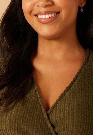 Femme souriante portant une boucle d'oreille en forme de cœur doré et un haut texturé vert olive à col en V, avec de longs cheveux foncés sur l'épaule.