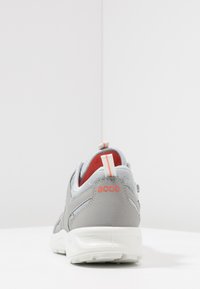 Sneaker in tessuto grigio con collare imbottito, suola bianca e dettagli arancioni, dotata di una linguetta sul retro e di un marchio sul tallone.