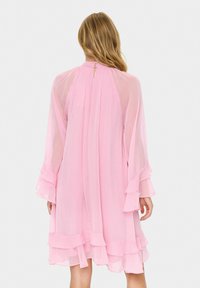 Rosa, luftiges Kleid mit langen, Glockenärmeln, geschichteten Rüschen am Saum und einer Schlüssellochöffnung am Rücken. Hergestellt aus leichtem, durchscheinendem Stoff.