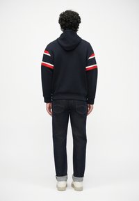 Sudadera azul marino con capucha y acentos de rayas rojas, blancas y negras en las mangas. Combinada con jeans de mezclilla oscura y zapatos beige.