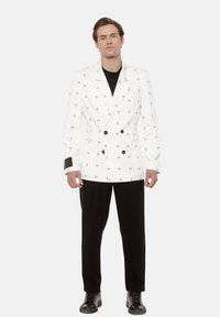 Mężczyzna stoi, ubrany w białą marynarkę podwójnie zapinaną z małymi czarnymi wzorami, czarną koszulę, czarne spodnie i czarne buty, na tle jednego koloru.