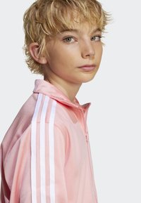 Rosa jeansjakke med glidelås laget av lett materiale; har hvite sidelister og en myk krage. Modellen har kort, uformell blondt hår.