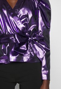 Haut cache-cœur métallique violet avec un nœud noué, présentant une texture lisse et un design structuré. Pantalon noir visible en bas.