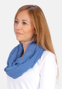 Écharpe bleue clair, enroulée, faite d'un tissu doux et texturé. Le design légèrement froissé ajoute de la dimension. Portée sur un haut blanc.