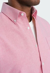 Vaaleanpunainen nappikauluspaita, jossa on klassinen kaulus, textuuripintainen kangas ja valkoiset napit, sekä vasemmalla puolella rintatasku.