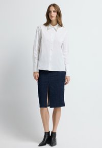 Witte blouse met lange mouwen en decoratieve kraag, gecombineerd met een marineblauwe kokerrok met een split aan de voorkant en een textuurpatroon. Zwarte enkellaarsjes.