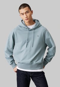 Felpa blu chiaro in tessuto morbido con grande tasca frontale, cappuccio con cordino e logo sul lato sinistro del petto. Abbinata a jeans scuri.