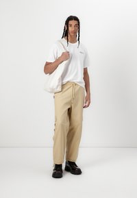 Hvit bomull t-skjorte med logo, beige cargo bukser med sidelommer og svarte støvler. Modellen bærer en kremfarget tote bag.