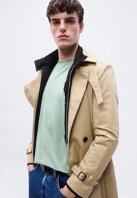 Νεαρός άνδρας με κοντά σγουρά μαλλιά, φορώντας μπεζ καμπαρντίνα πάνω από ανοιχτό πράσινο πουκάμισο και μπλε τζιν, με τα χέρια στις τσέπες, ουδέτερο φόντο.