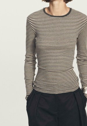 Personne portant une chemise ajustée à manches longues beige et noire rayée, rentrée dans un pantalon noir taille haute, les mains dans les poches.