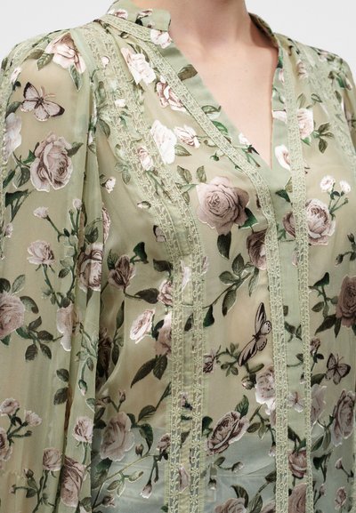 Blouse légère vert clair avec motifs fleurs roses et papillons ainsi que des bordures en dentelle verticale sur le devant et les manches.