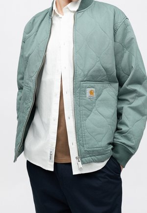 Person iført en lysegrøn quiltet jakke over en hvid skjorte med knapper og en brun undertrøje, med mørkeblå bukser og hænderne i lommerne.