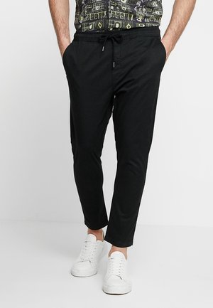Man in zwarte joggingsbroek met zijzakken en witte sneakers, staand tegen een plain witte achtergrond.