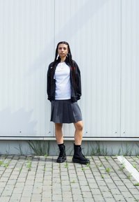 Černá zipová bunda s texturovaným materiálem, pod ní bílý tričko, šedá plisovaná sukně a černé bojové boty. Pozadí tvoří vertikální kovové panely.