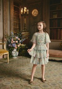 Egy réteges, fodros ruha pasztell virágmintával és rövid ujjakkal. A modell bronz Mary Jane cipőt visel, és egy mintás szőnyegen áll.