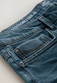 Jeans en denim d'une teinte bleu moyen avec une texture lisse, dotés d'une poche frontale, de coutures contrastées et d'une étiquette de marque visible au niveau de la taille.