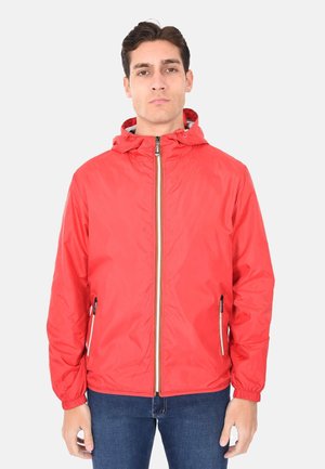 Giovane uomo in piedi di fronte, indossa una giacca con cappuccio rossa con zip e jeans blu su sfondo chiaro uniforme.