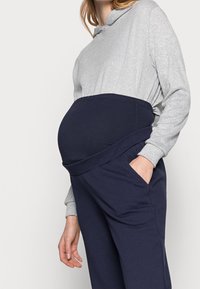 Těhotenský outfit obsahující šedou mikinu s dlouhým rukávem a tmavě modré kalhoty s širokým pasem, navržený pro pohodlí a podporu.