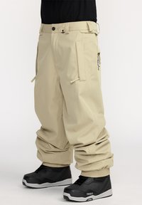 Beige cargobroek met een ontspannen pasvorm, voorzien van meerdere zijzakken, verstelbare enkelboorden en gedragen met zwarte sneakers.