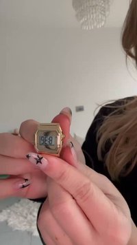 Montre-bracelet numérique dorée avec un boîtier rectangulaire, un écran LCD affichant l'heure et un design de surface texturé. Ongles décorés d'un motif étoilé dans le cadre.
