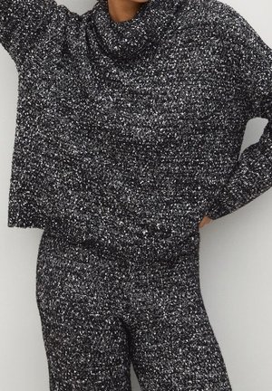Femme portant un pull en maille texturée noir et blanc à épaules tombantes et un pantalon en maille assorti, posant avec une main sur la hanche.