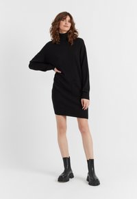 Schwarzes gestricktes Kleid mit hohem Kragen, langen Ärmeln und zwei Seitentaschen; kombiniert mit schwarzen klobigen Knöchelstiefeln.