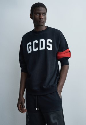 Ενήλικος άνδρας που φοράει μαύρο φούτερ GCDS με κόκκινη ρίγα στο μανίκι και μαύρο παντελόνι που ταιριάζει, σε γκρι απλό φόντο.