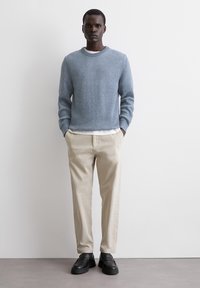 Hellblaues, strukturiertes Strickpullover mit Rundhalsausschnitt; cremefarbene Hose; schwarze Slipper; stehende Figur mit Händen in den Taschen.