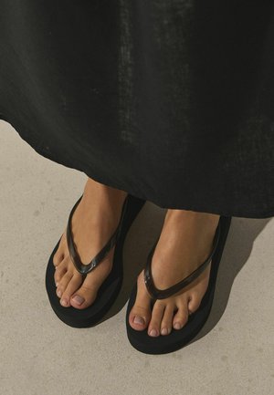 Feet wearing black flip-flops with glossy straps, partially covered by a long black garment, standing on a light-colored floor.
