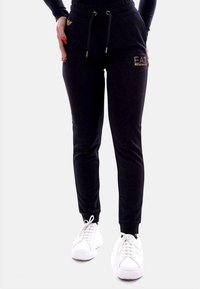Zwarte joggingsbroek met een elastische tailleband, koord, zijzakken en gouden logo-accenten, gecombineerd met witte sportschoenen.
