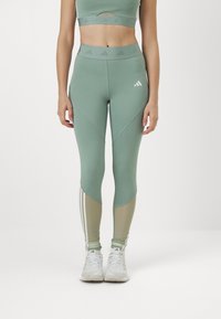 Grønne atletiske leggings med høy midje, kontrasterende beige paneler og tre hvite striper langs de nedre leggene.