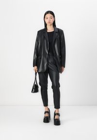 Zwarte leren blazer gecombineerd met bijpassende slim-fit broek. Model houdt een kleine zwarte handtas vast. Schoenen hebben dikke zolen en zijkleurige accenten.