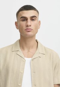 Camisa de manga corta a rayas beige con cierre de botones en el frente y una textura suave, llevada sobre una camiseta sin mangas blanca, exhibida sobre un fondo neutro.