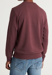 Czerwony sweter z długim rękawem o rypsowej fakturze i okrągłym dekolcie, z prostym wykończeniem i metką po lewej stronie. W zestawie z niebieskimi dżinsami.